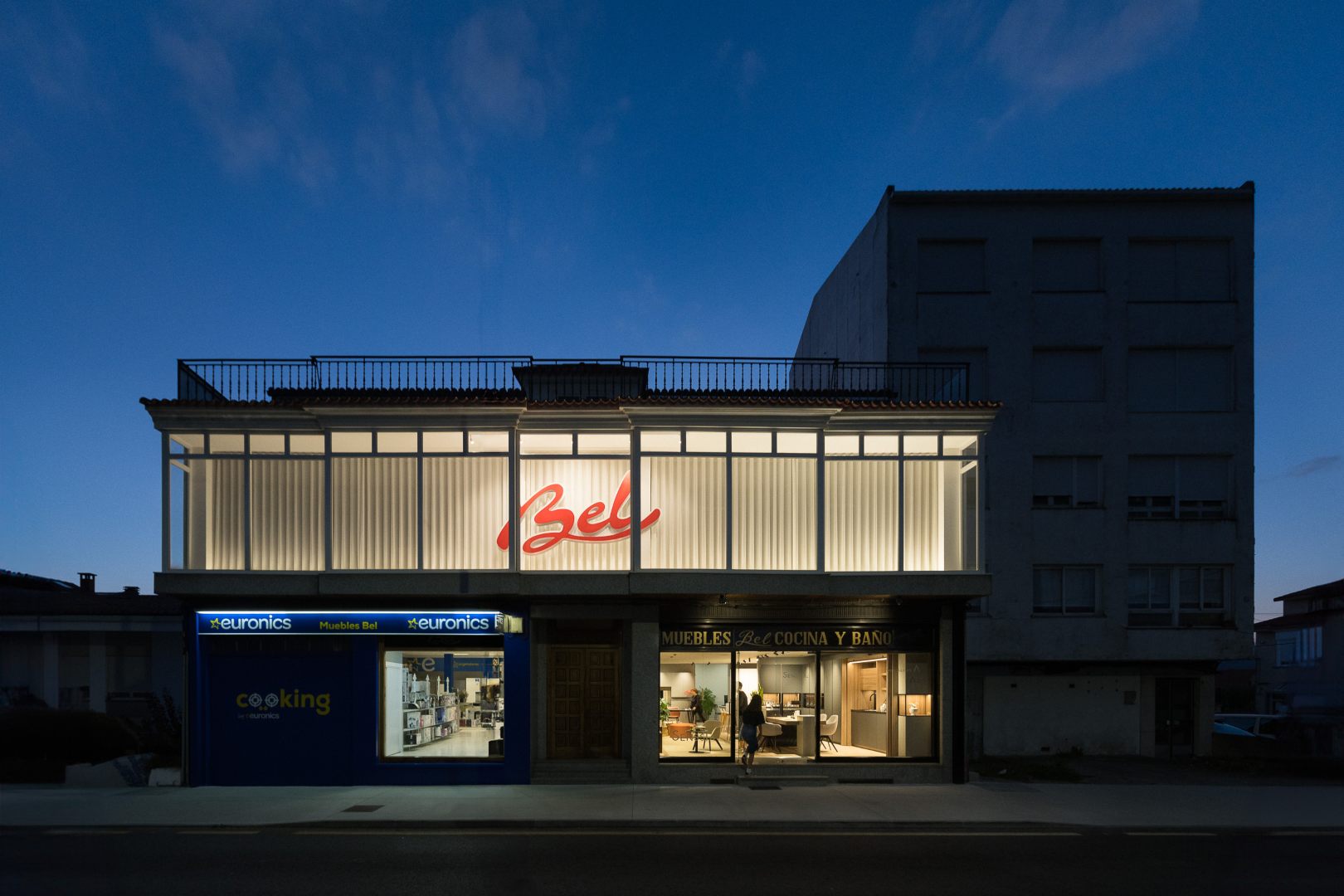 Fachada de la tienda Muebles Bel por la noche, iluminada con el letrero encendido