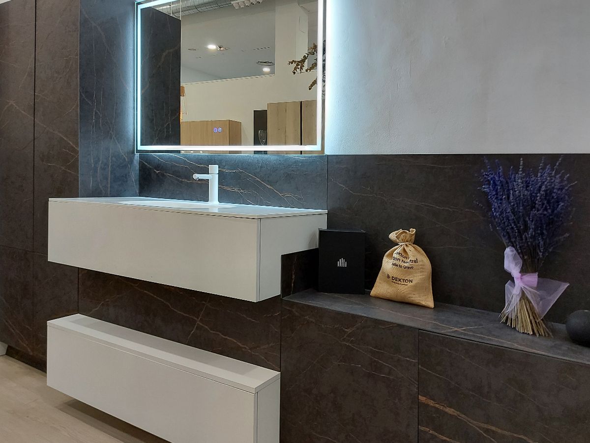 Composición moderna para baño en exposición con lavabo en blanco, muebles en acabado cerámica oscura y espejo rectangular con leds