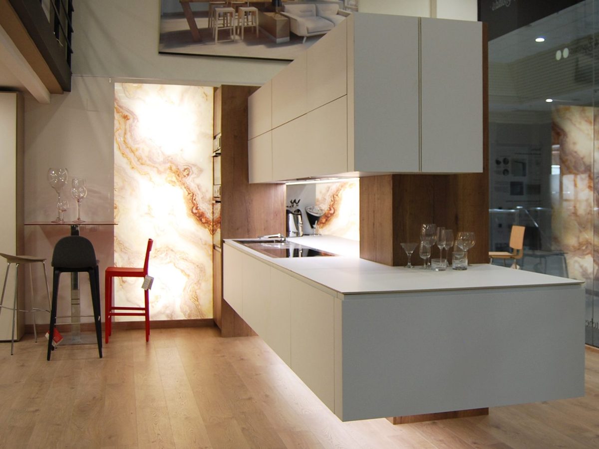 Cocina en península con lateral en "U" en exposición con mobiliario combinado en blanco y madera, mesa alta con sillas y pared iluminada imitando al mármol